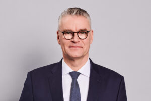 Portraitfoto von Dr. Constantin H. Alsheimer. Herr Alsheimer hat kurze graue Haare, keinen Bart und trägt Brille. Er trägt ein weißes Hemd, dunkles Jackett, Krawatte.