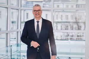 Portraitfoto von Dr. Alsheimer. Herr Alsheimer trägt einen dunklen Anzug, eine Krawatte und steht vor Fenstern.